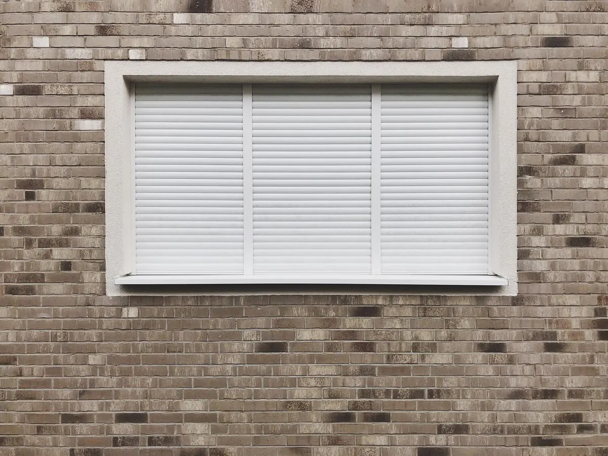 Fenêtre à trois vantaux fermée par des volets roulants blancs dans une façade en brique, photo d'illustration